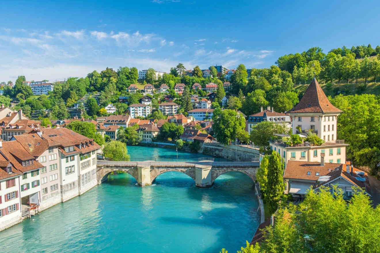 View of a picturesque European town with a stone bridge crossing a turquoise river, surrounded by greenery and traditional buildings under a clear blue sky.