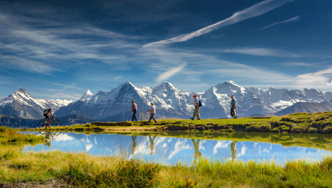 A group of people walk and bike along a grassy path beside a reflective lake, with snow-capped mountains in the background under a blue sky.