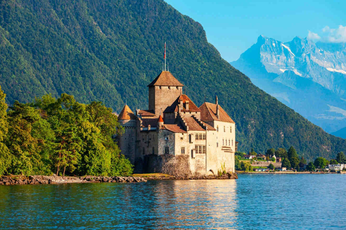 A historic castle with red-roofed turret sits on the edge of a lake, flanked by lush greenery and backed by tall, rugged mountains under a clear blue sky.