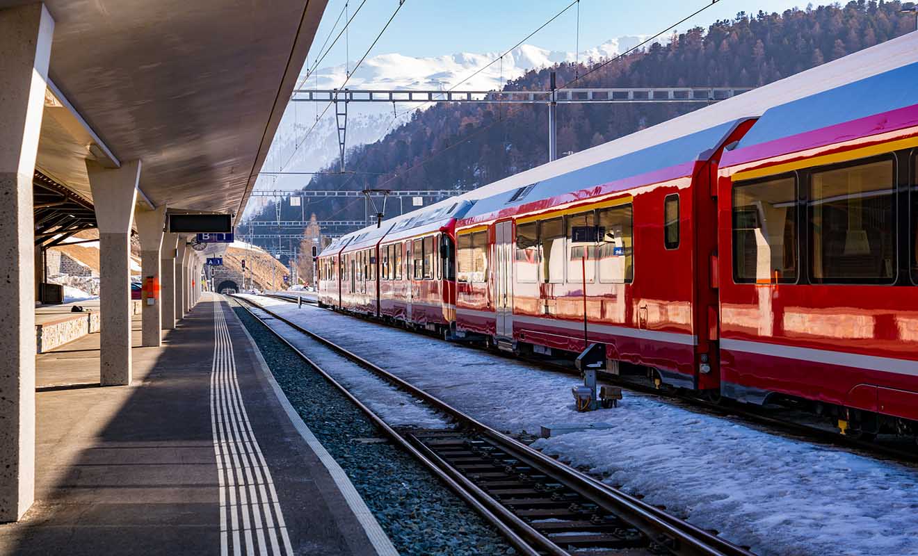 A red passenger train is stationed on snow-covered tracks at a mountain railway station on a sunny day, with snow-capped mountains in the background.