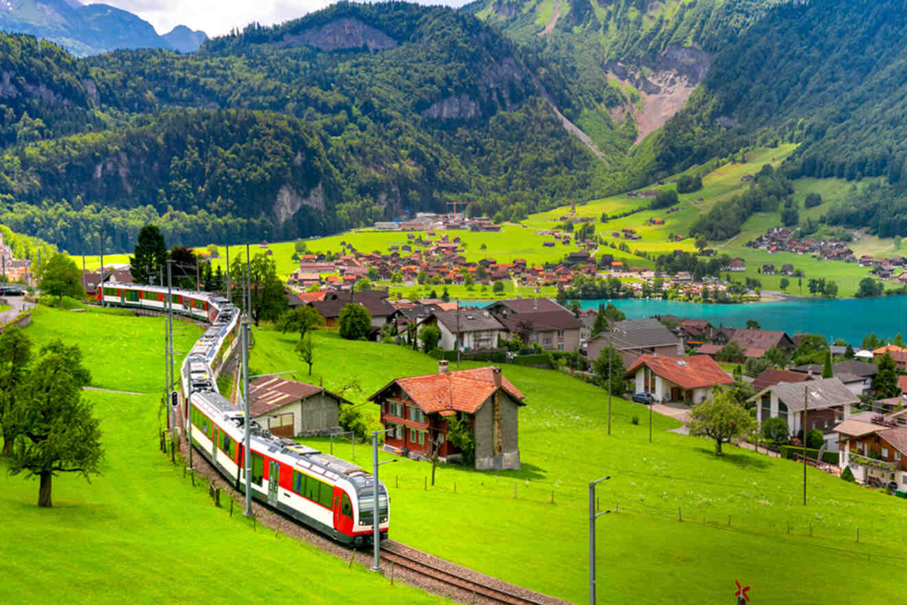 A red and white train passes through a scenic rural landscape with green fields, scattered houses, a small lake, and forested mountains in the background.