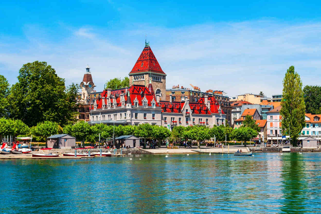 A scenic view of a lakeside town with a prominent building featuring a red roof and turret, surrounded by greenery and water in the foreground.