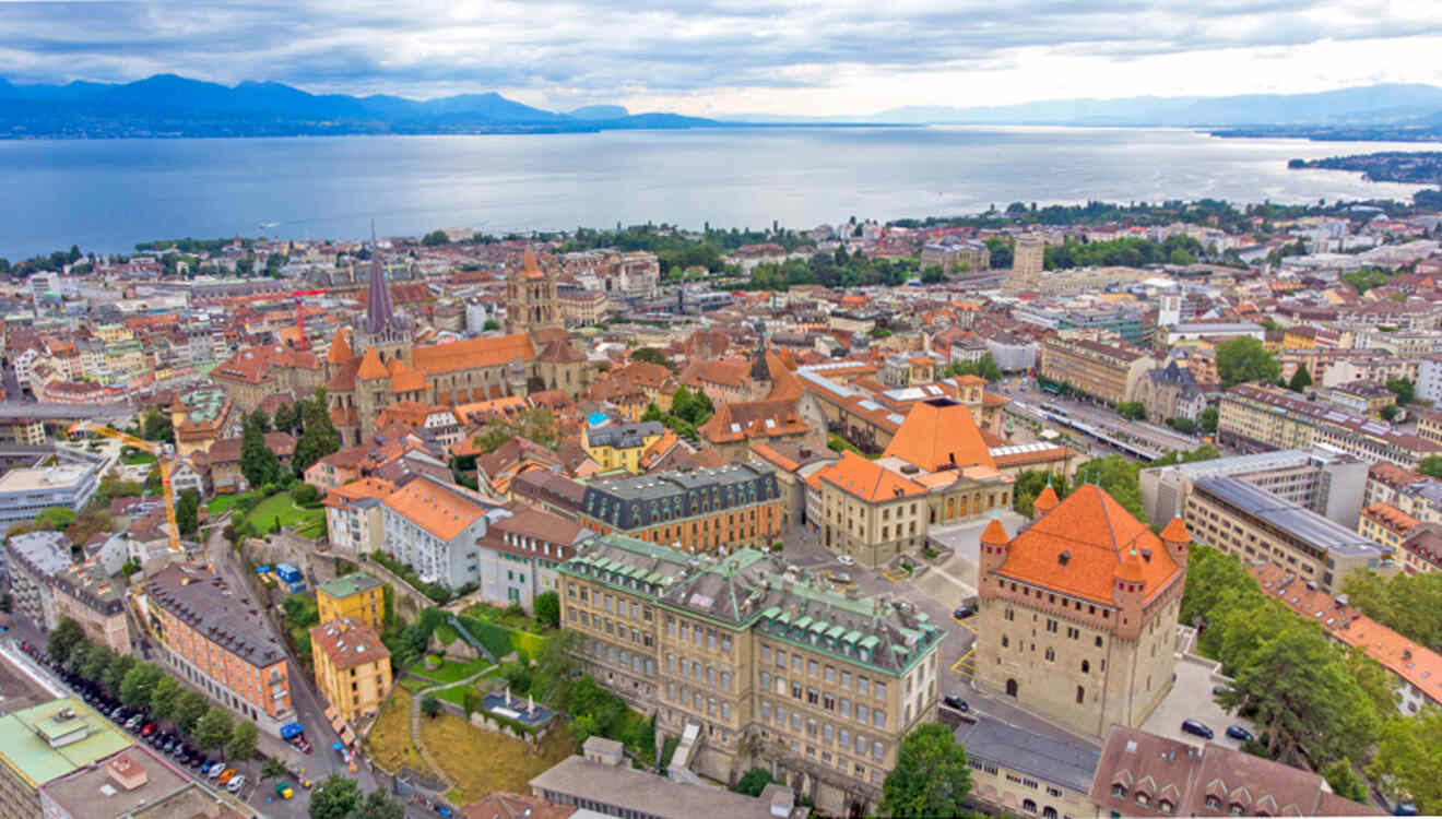 Aerial view of a European city with numerous buildings and a cathedral, situated near a large lake with mountains in the distance.