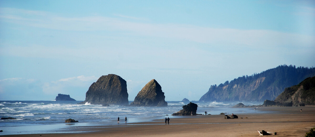 Wide beach with people walking on the sand, ocean waves, and large rock formations offshore beneath a hazy blue sky with distant wooded hills.