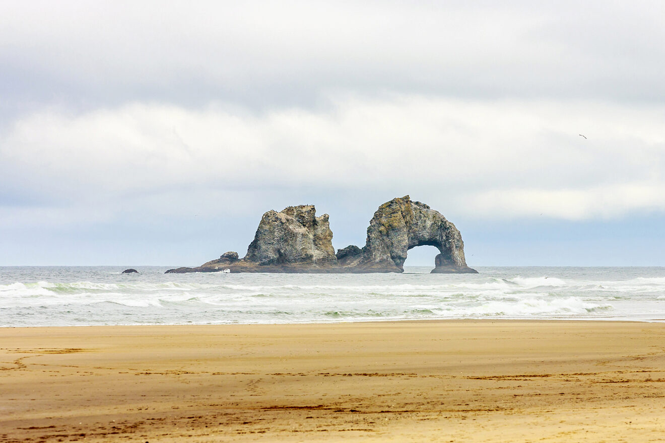 A coastal rock formation with an arch in the middle, surrounded by ocean waves, viewed from a sandy beach under a cloudy sky.