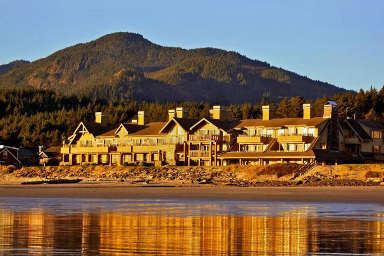 A large, multi-story lodge with numerous windows and balconies stands by the waterfront with forested hills in the background, reflecting on the calm water.