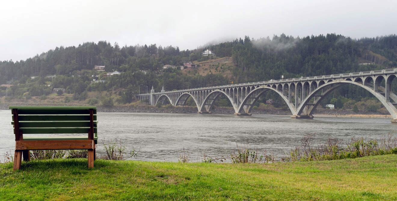 A green bench overlooks a river with a large arch bridge in the background and forested hills on a foggy day.