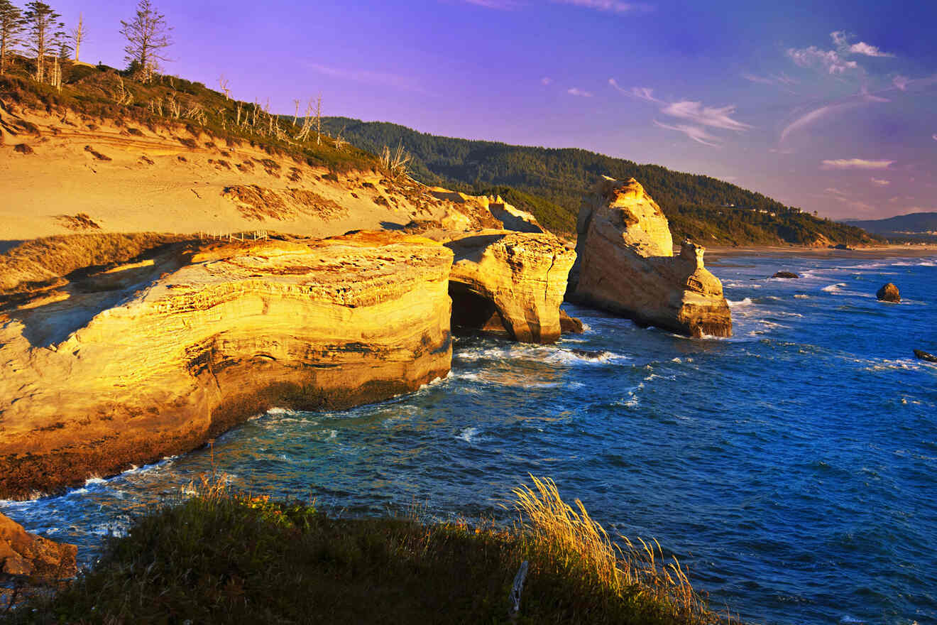 A coastal landscape with rugged cliffs, waves crashing on rocks, sandy dunes, and a mountainous backdrop under a clear sky.