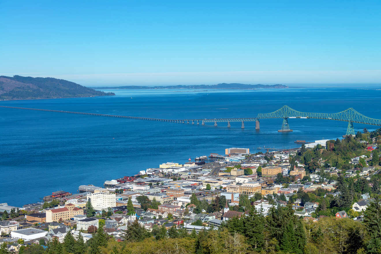 Aerial view of a coastal town featuring a long bridge extending over a large body of water with mountainous terrain visible in the background under a clear, blue sky.