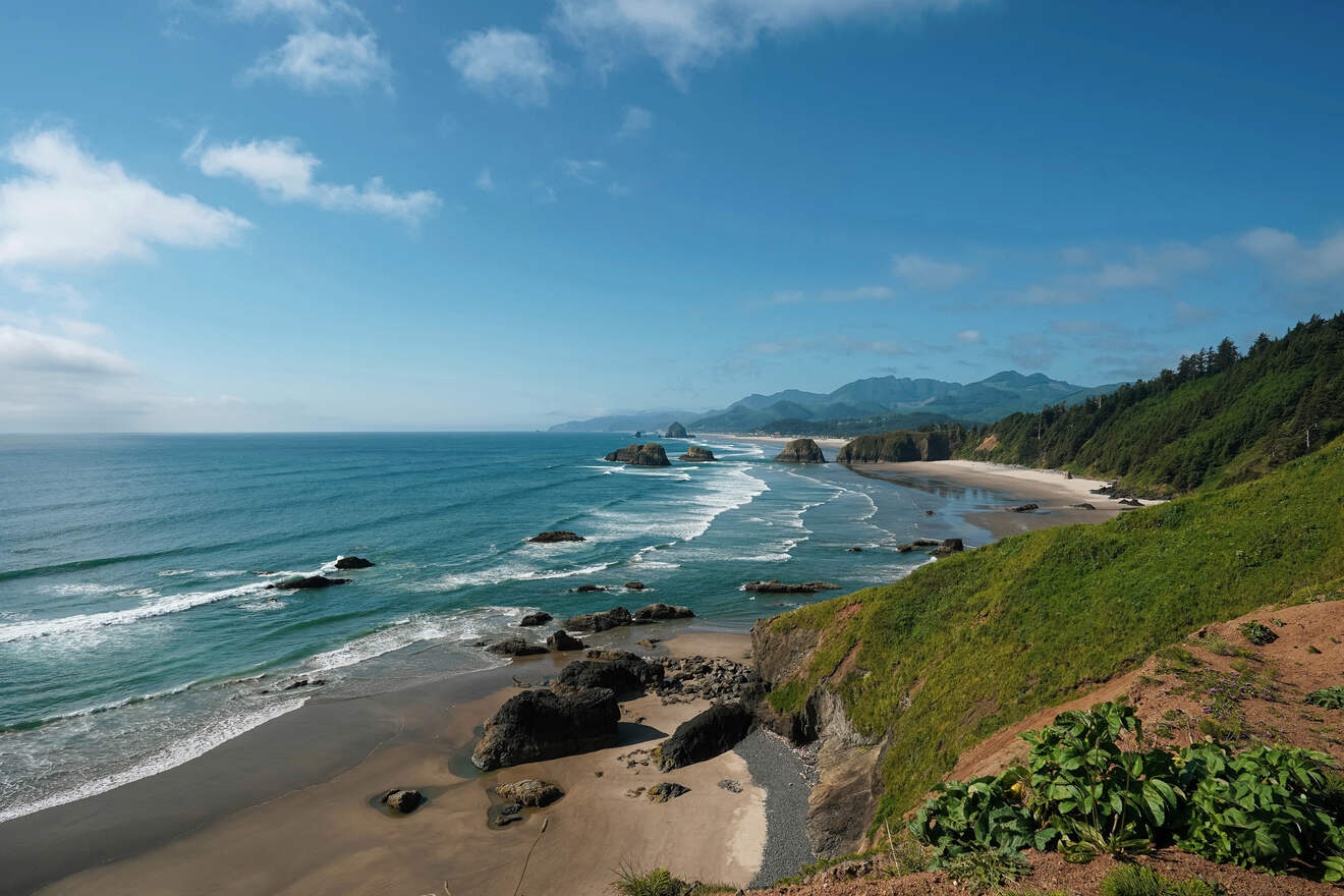 A scenic coastal view of a rocky shoreline with waves gently crashing against the beach, under a clear blue sky. Hills covered in greenery stretch alongside the coast, and mountains are visible in the distance.