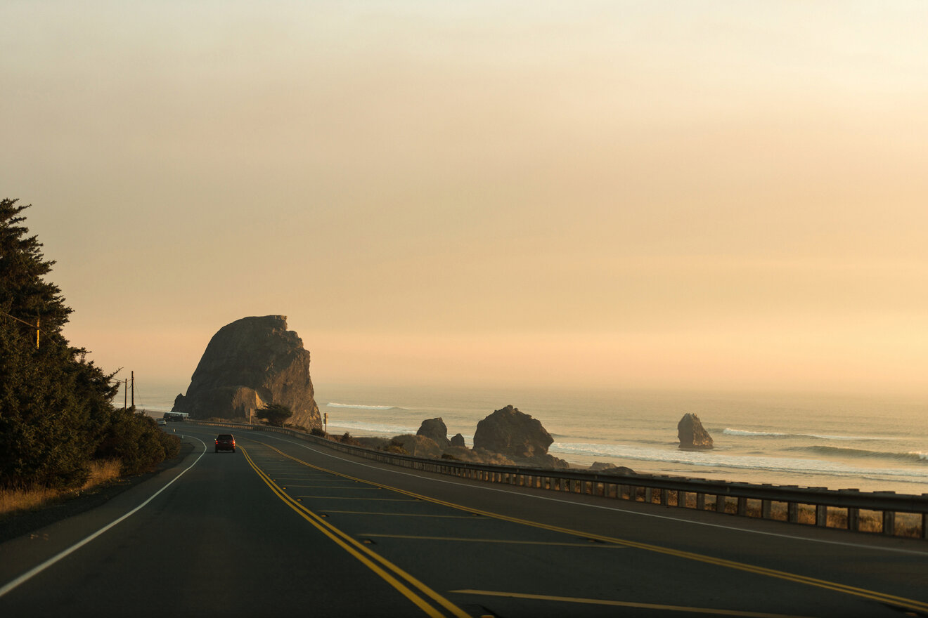 Two-lane road winding along a coastal landscape with large rocks and a calm ocean under a hazy sky, with a car driving in the distance.