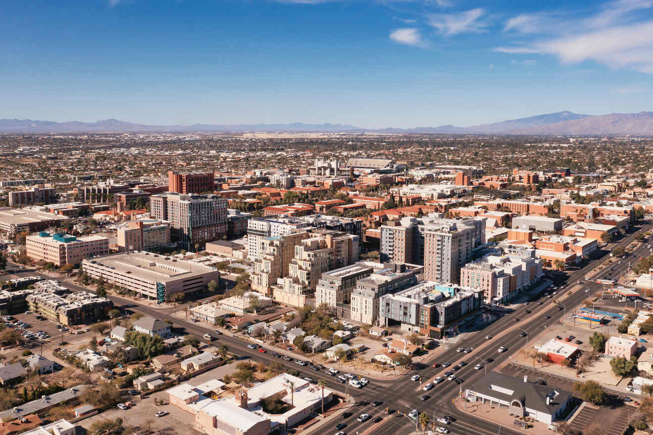 Aerial view of a cityscape with numerous buildings, streets, and vehicles, set against a backdrop of mountains under a blue sky with scattered clouds.