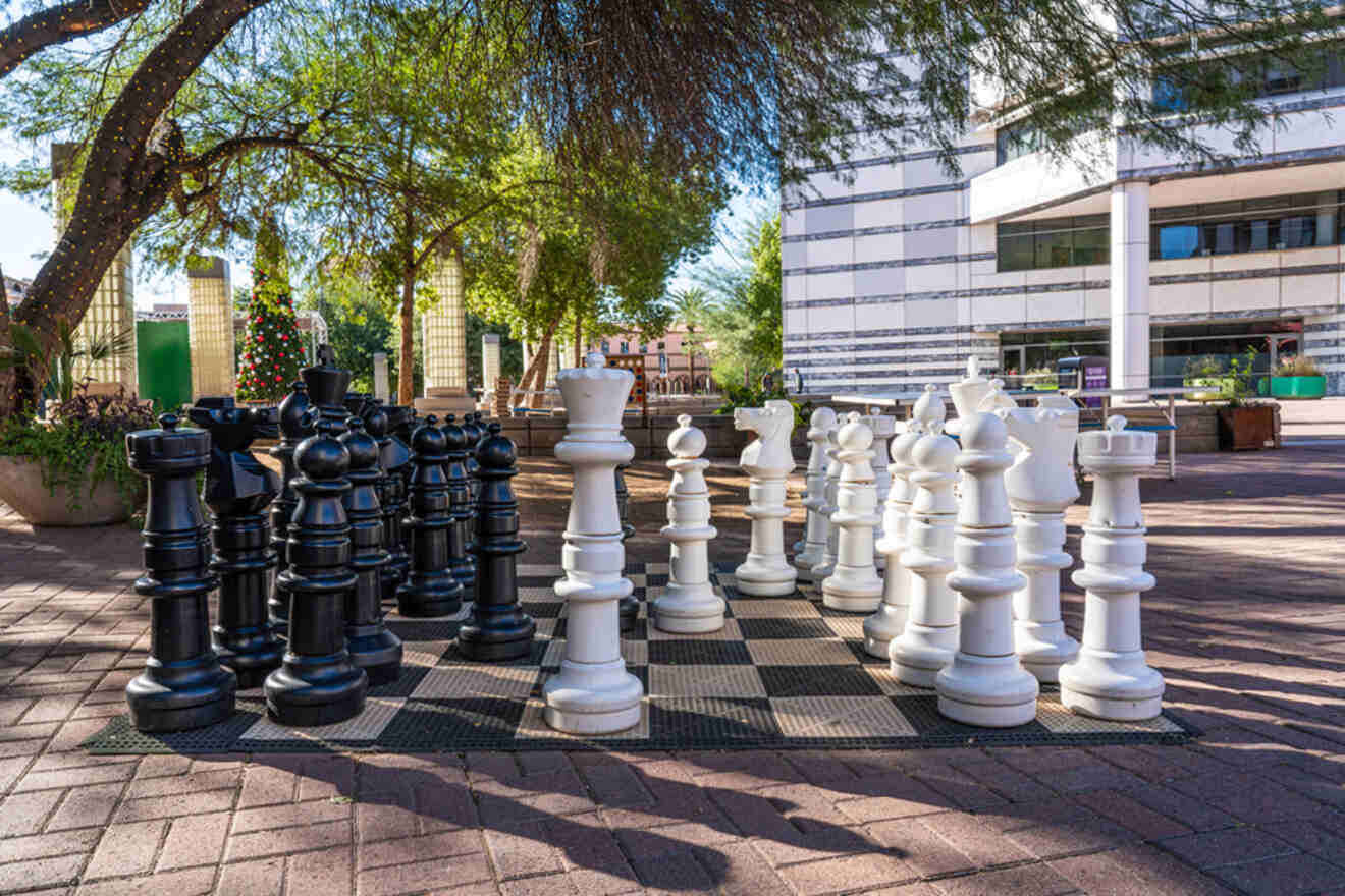 A large outdoor chess set with black and white pieces is arranged on a chessboard. The setting is a sunny plaza with trees and a modern building in the background.