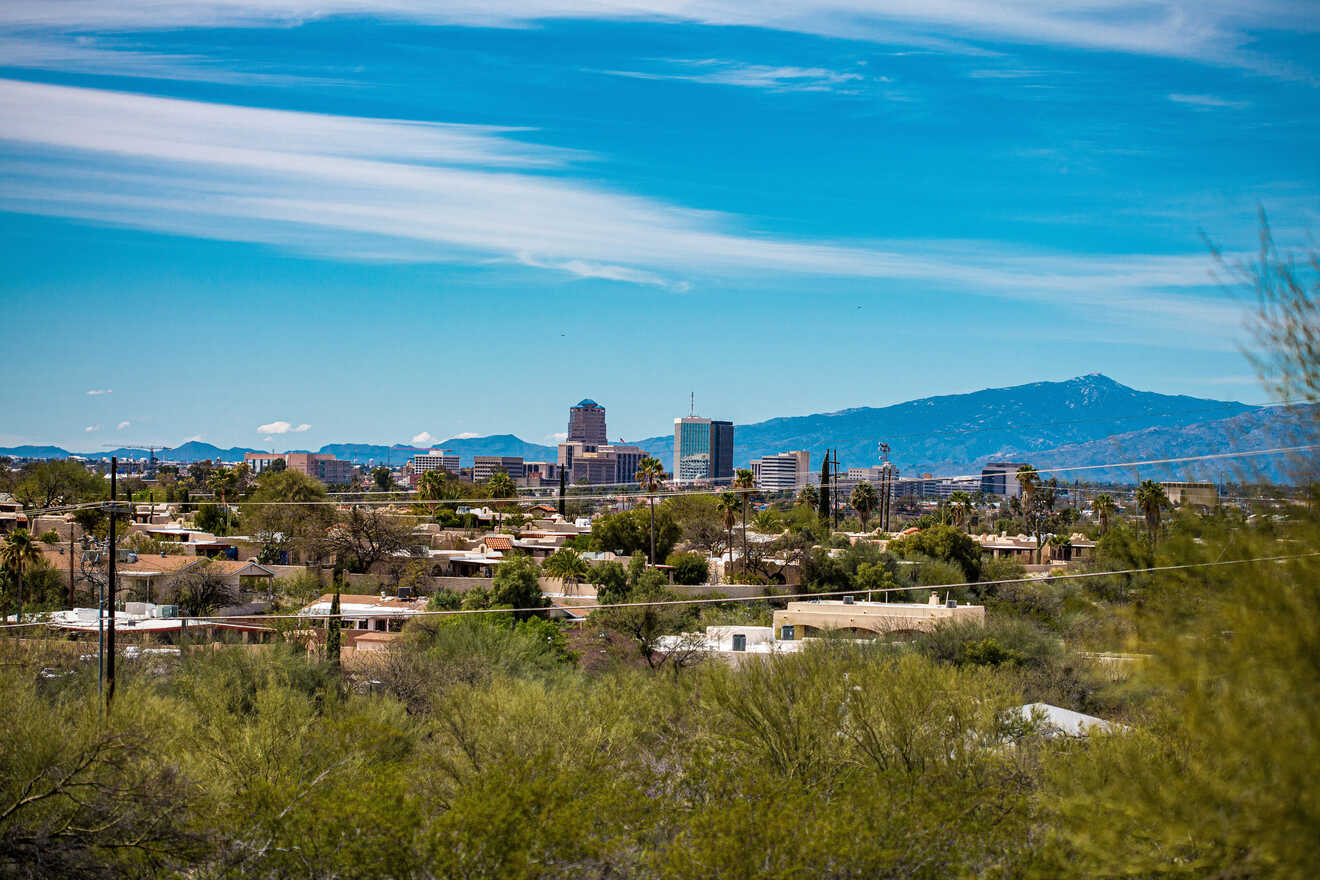 A distant view of a cityscape with a mixture of tall buildings and residential areas in front, set against a backdrop of mountains and a blue sky with scattered clouds.