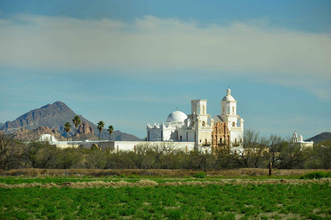 A historic white mission church with domes and bell towers stands against a backdrop of mountains and clear sky, surrounded by desert vegetation and green fields.