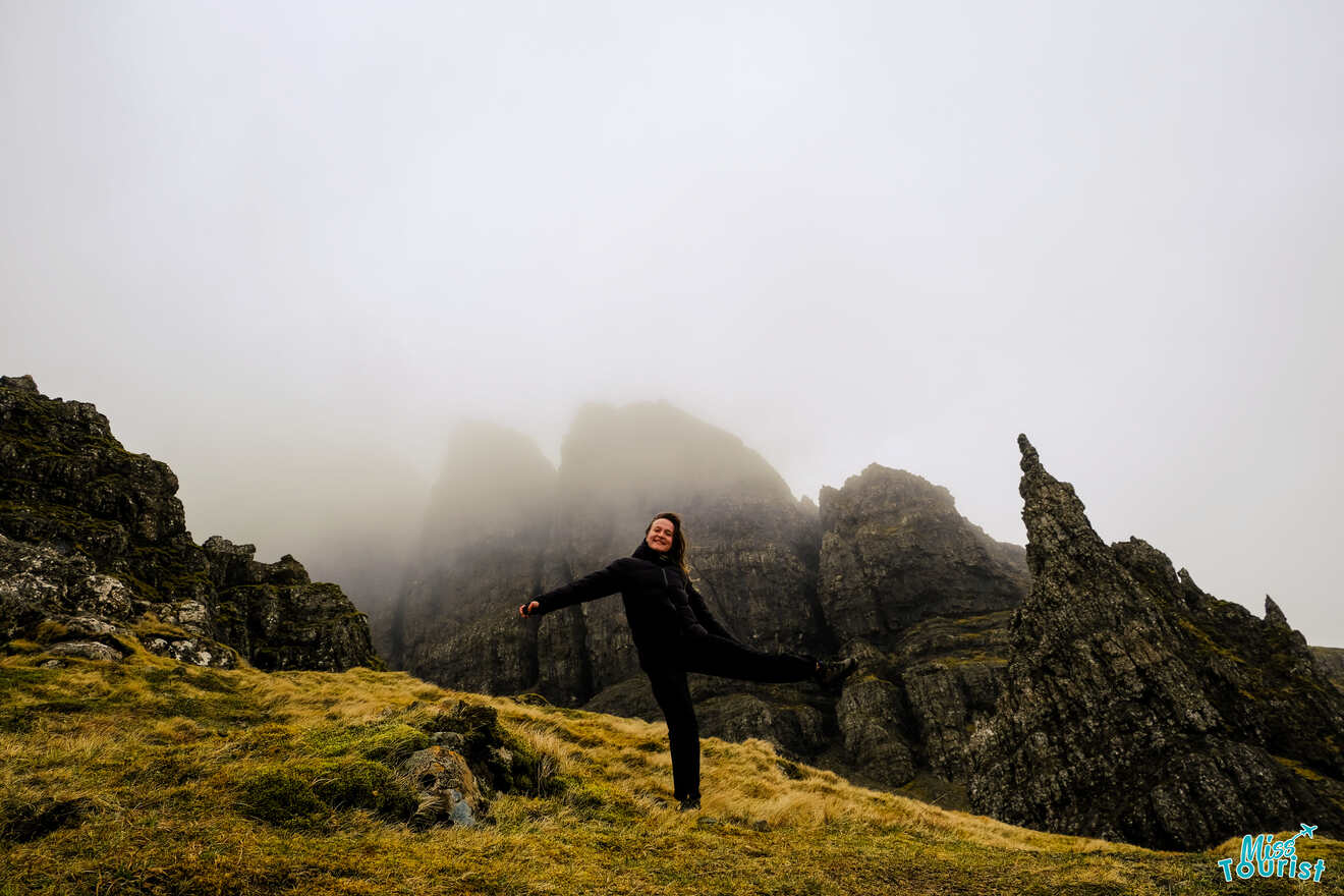 The writer of the post standing on a grassy slope, striking a playful pose with one leg lifted, surrounded by rocky formations and misty, cloud-covered peaks.