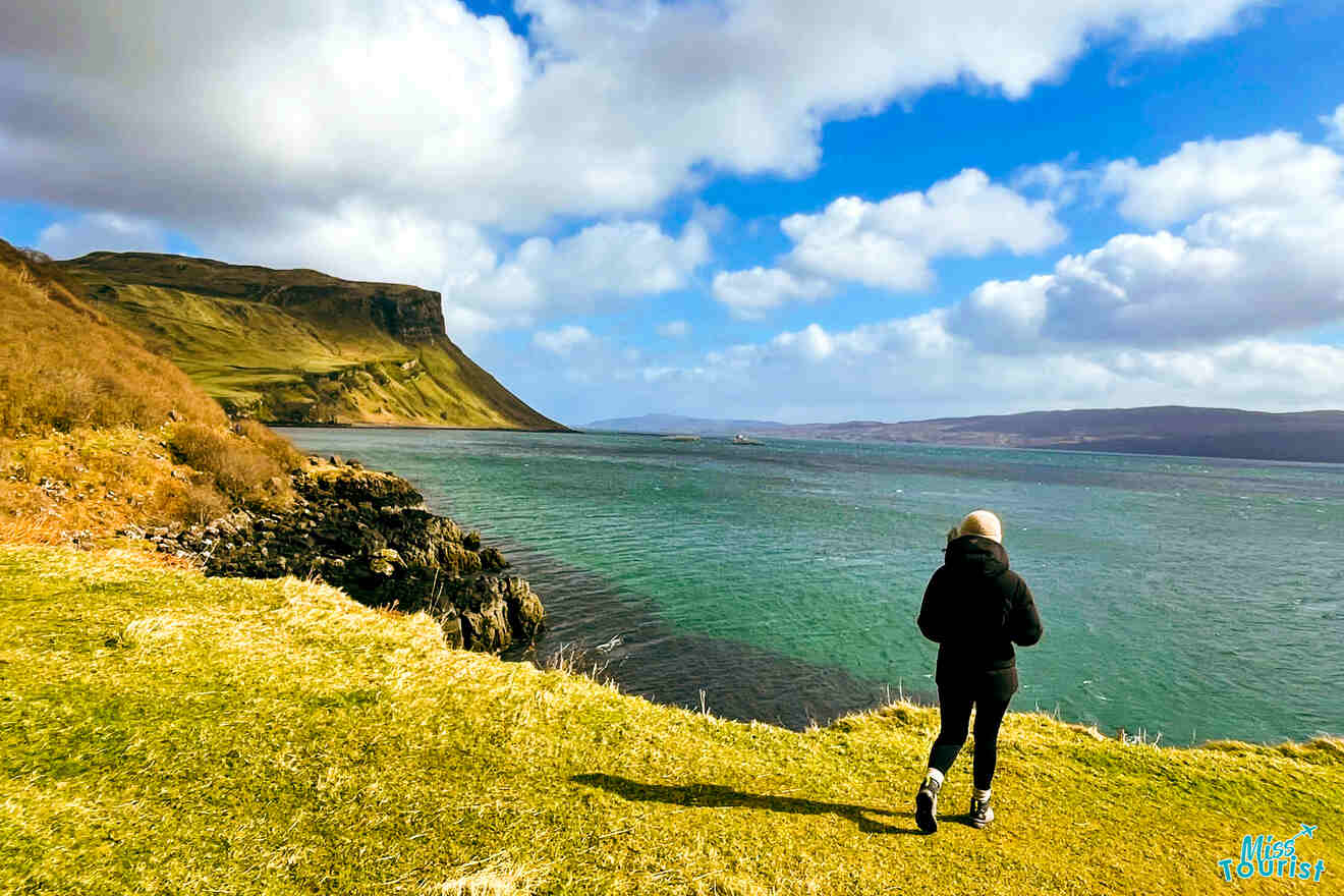 The writer of the post in a black jacket and hat walks near a grassy coastline overlooking a body of water with hills and cliffs in the distance under a partly cloudy sky.