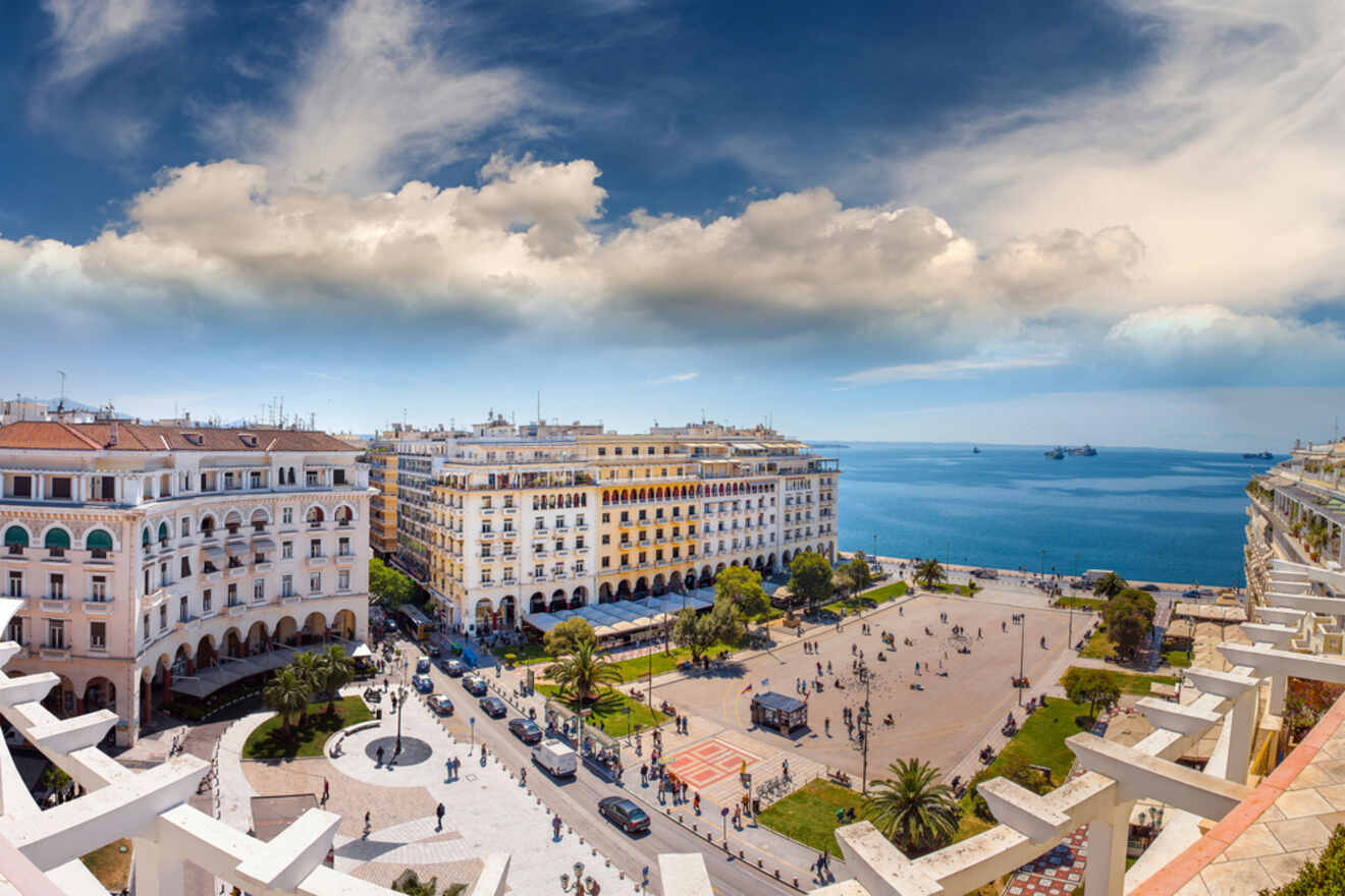 A coastal cityscape featuring a large public square surrounded by historic buildings, with the ocean and several ships visible in the background under a blue sky with scattered clouds.