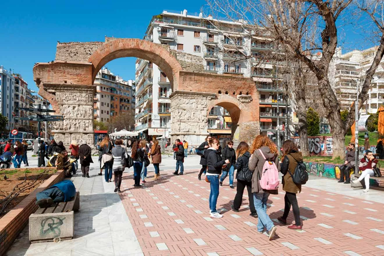 People walk near the Arch of Galerius in a busy urban area, surrounded by modern buildings and trees on a sunny day.