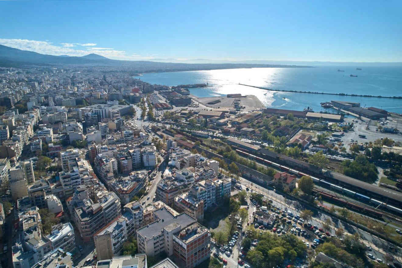 Aerial view of a coastal city with densely packed buildings, a busy port, and a visible shoreline under a clear blue sky.