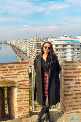 the author of the post in a black coat and sunglasses stands on a brick balcony overlooking a cityscape with a coastal view.