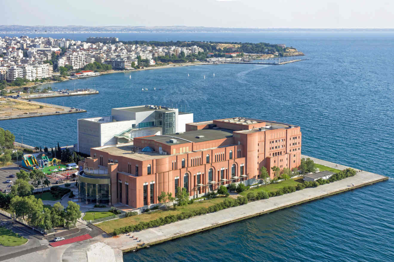 Aerial view of a large building by the sea, with a cityscape in the background and a waterfront promenade surrounding the structure.