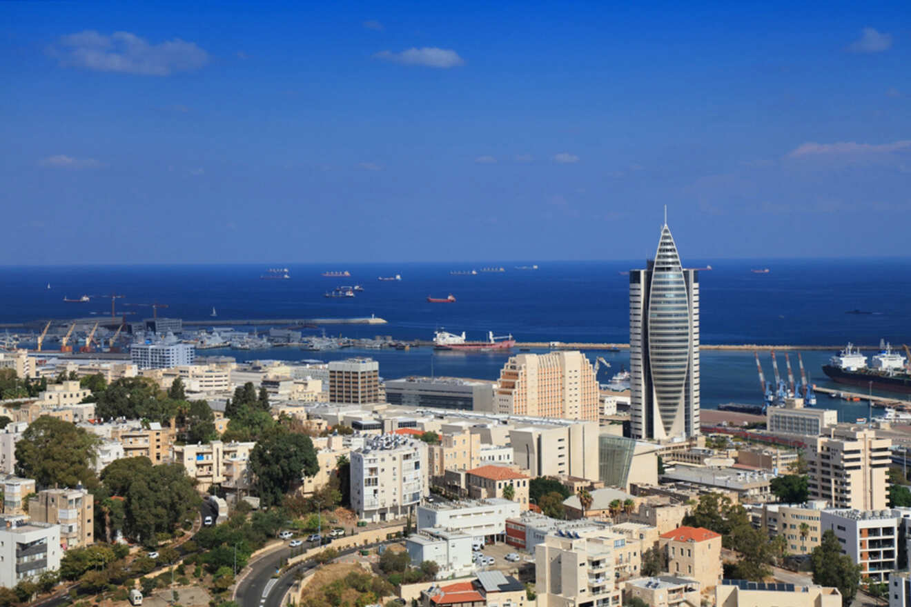 Aerial view of a coastal city with modern buildings, a prominent tower, and ships in the sea under a clear blue sky.