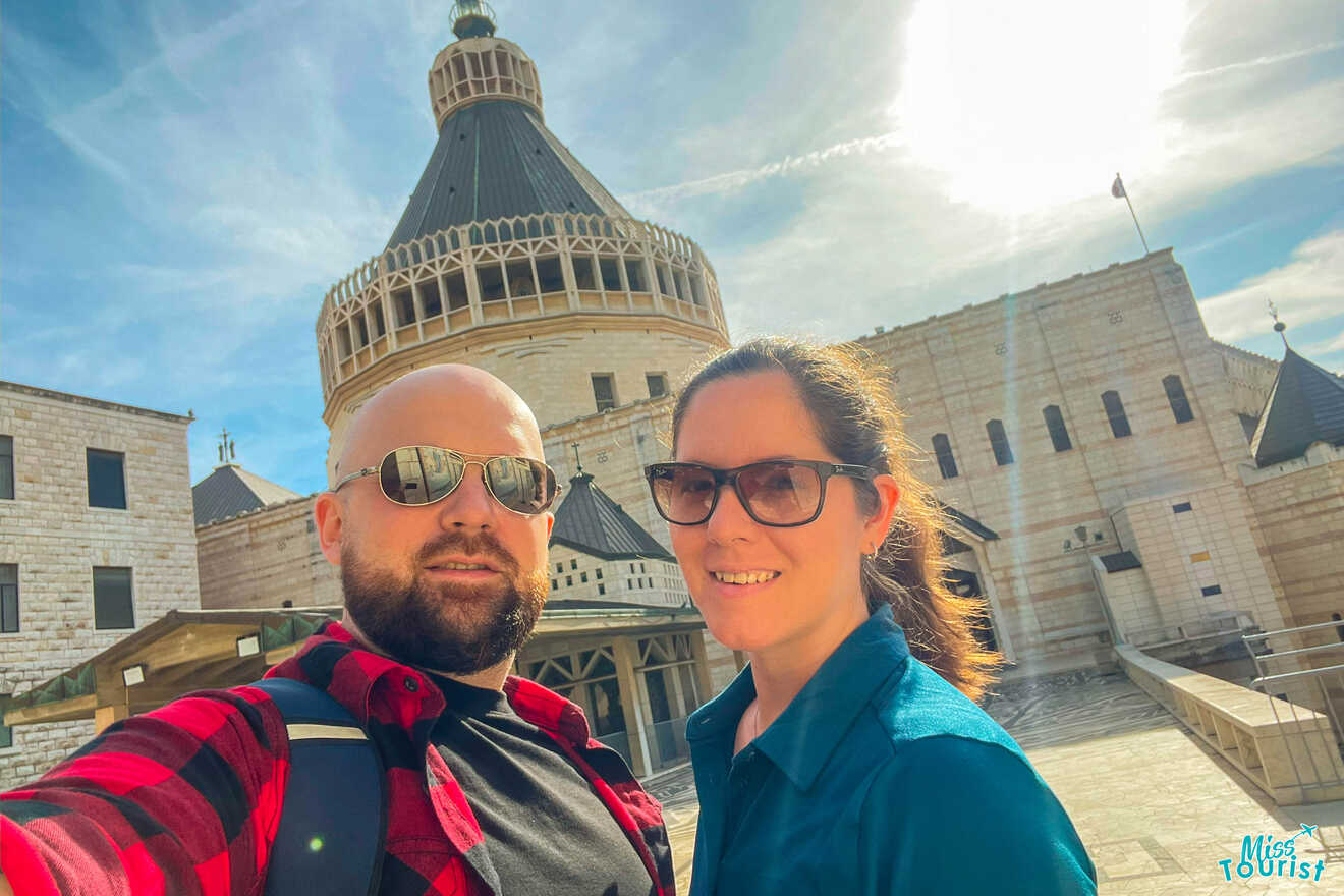 The writer of the post with her husband taking a selfie in front of the Basilica of the Annunciation in Nazareth with a sunny sky in the background.