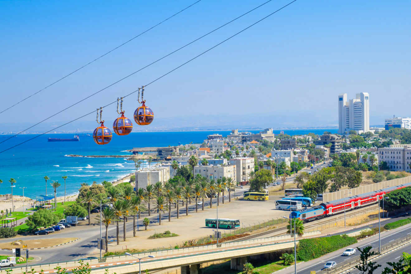 Cable cars in Haifa, Israel, glide above a coastal cityscape with buildings, a train, and the sea in the background on a clear day.