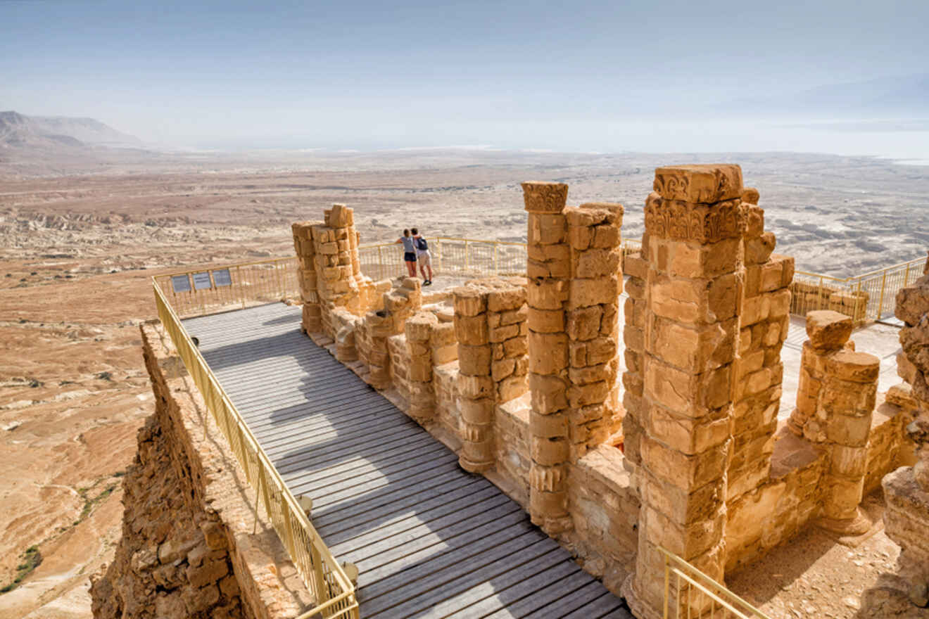 Two people stand on a walkway among ancient stone ruins, overlooking a vast desert landscape under a clear sky.