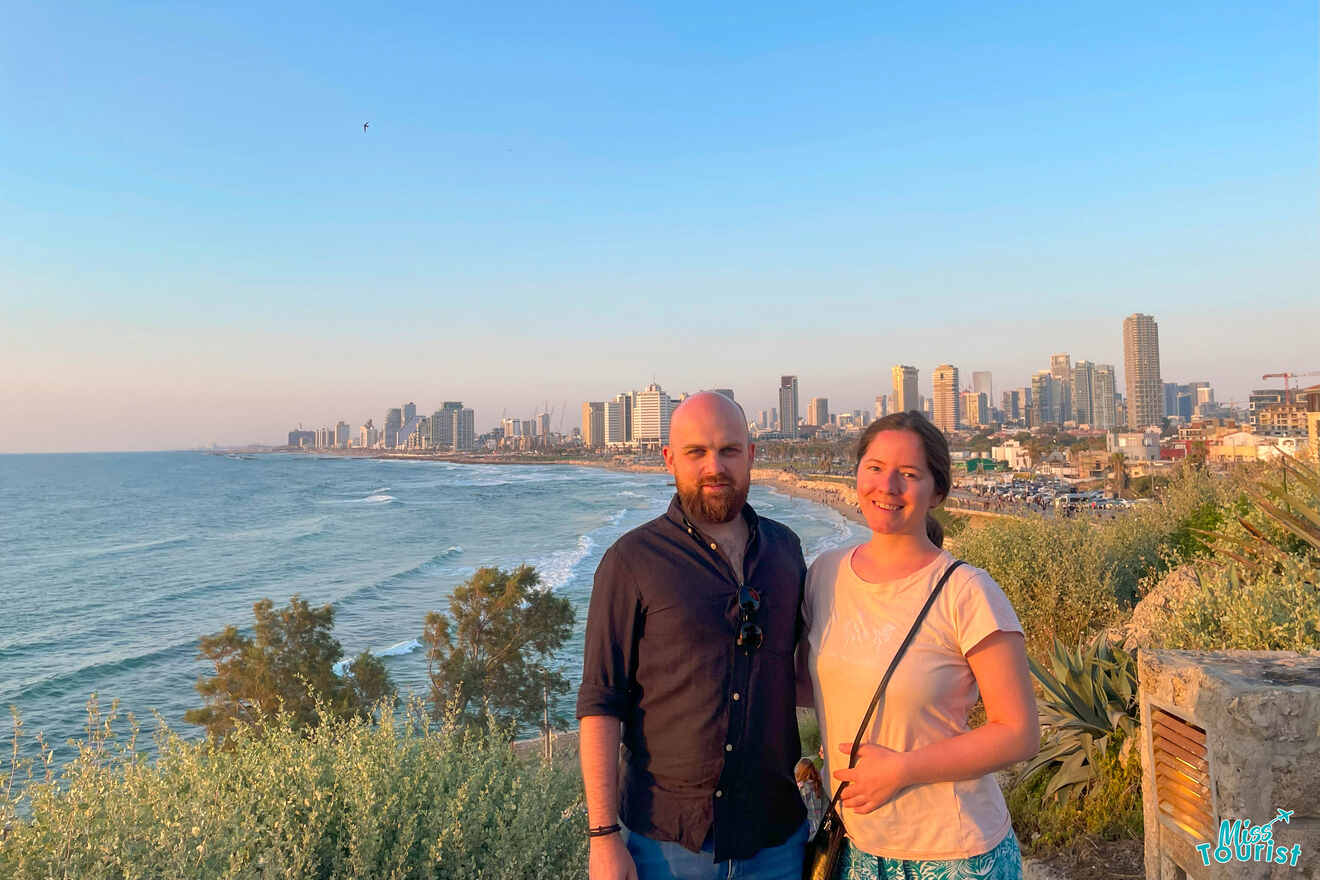 The writer of the post with her husband stands on a cliff overlooking a coastal cityscape with skyscrapers and the sea at sunset.