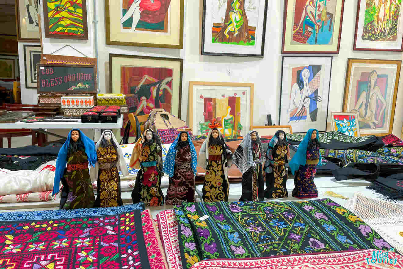 Various colorful textiles and patterned dolls are displayed on a table in front of a wall filled with framed artworks.