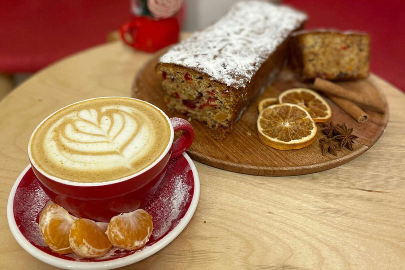 A red cup of latte with foam art sits beside a wooden board with sliced cake, dried oranges, and star anise on a light wooden table.