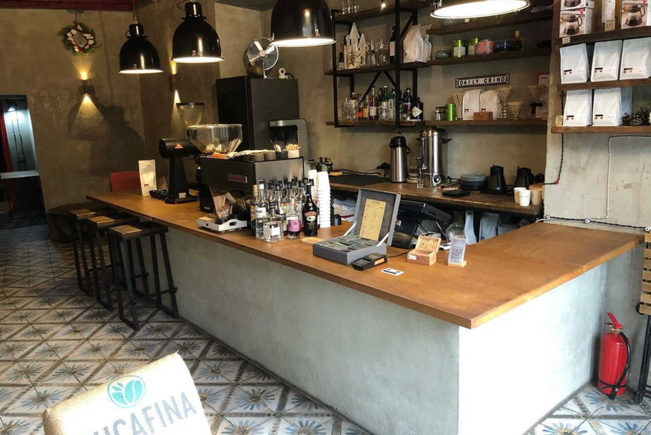 Coffee shop interior with a long wooden counter, bar stools, espresso machines, and shelves with various items, including coffee equipment and decor. Tile flooring with geometric pattern.