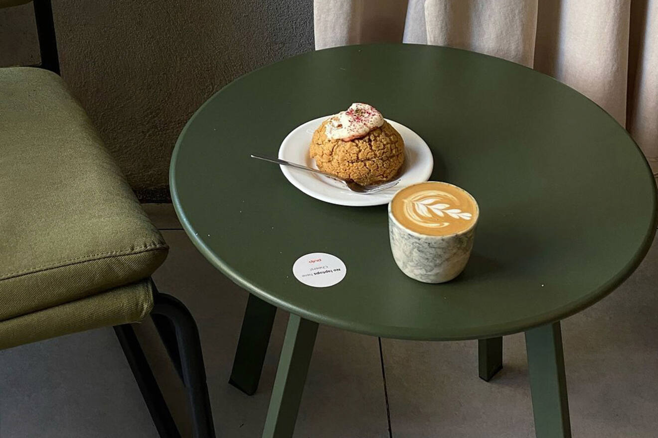 A green table with a latte in a marble-patterned cup and a cream-filled pastry on a white plate. Nearby is a green cushioned chair.