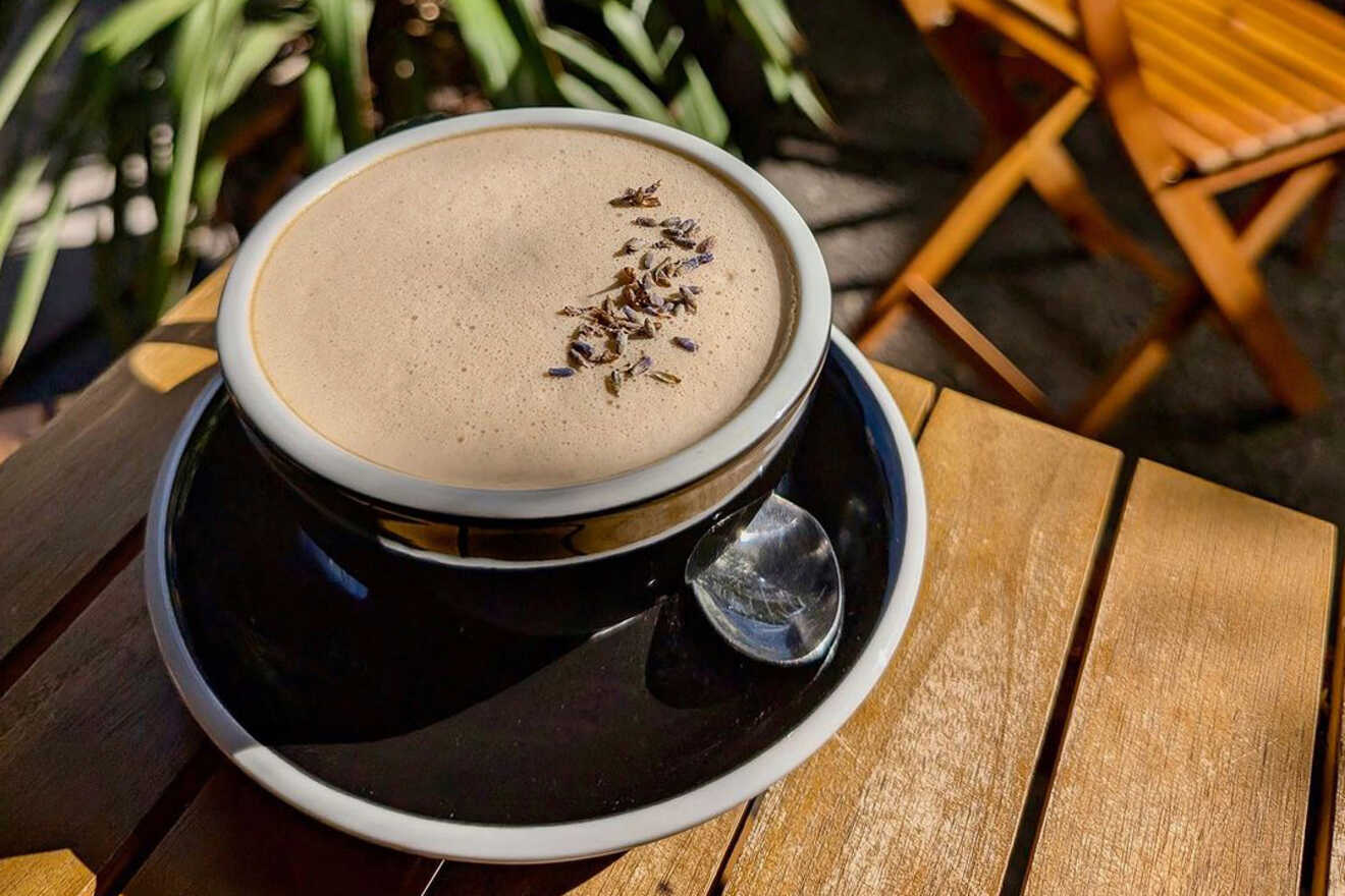 A cup of frothy coffee topped with dried lavender sits on a black saucer with a spoon, placed on a wooden table.