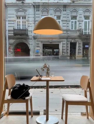 Two wooden chairs and a table with flowers, a pen, and a glass inside a café window with a view of a blurred street and historic building.