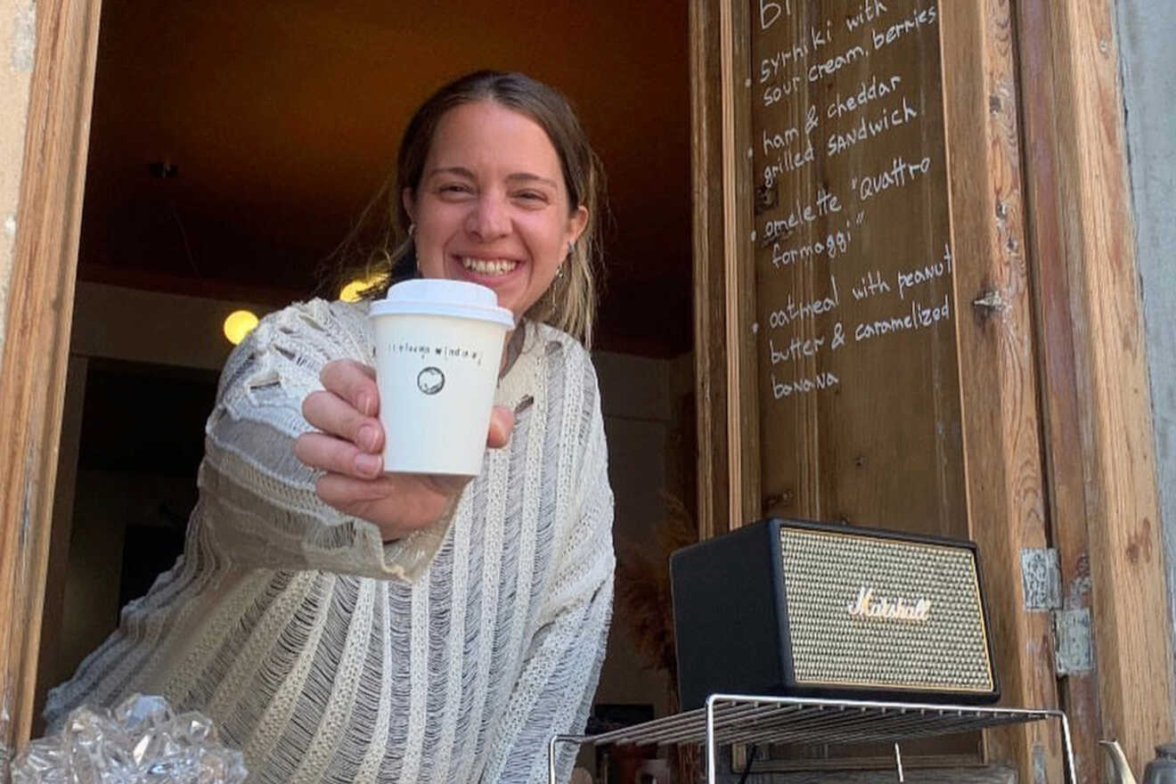 A person in a striped sweater holds out a coffee cup from a wooden window serving area. A menu is written on the wall.