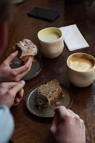 Two people enjoying pastries and drinks at a table. One holds a cinnamon roll, the other has a fork over a slice of bread. Two cups of coffee and a phone are also on the table.