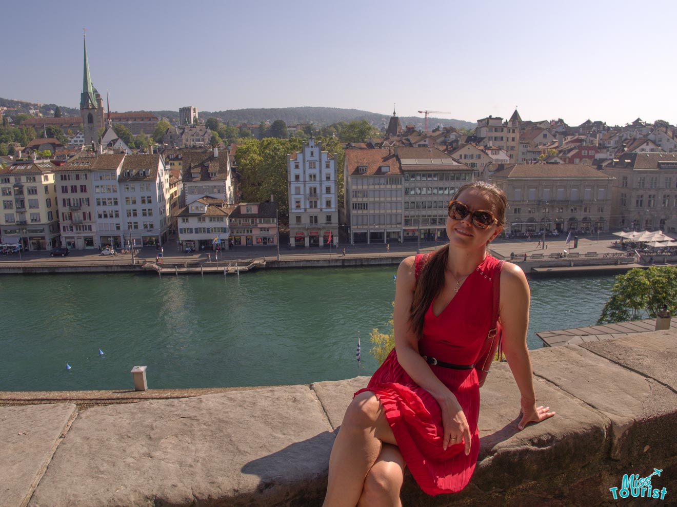 Yulia, the founder of this website, in a red dress and sunglasses sits on a stone ledge overlooking a river and a town with buildings and a church in the background.