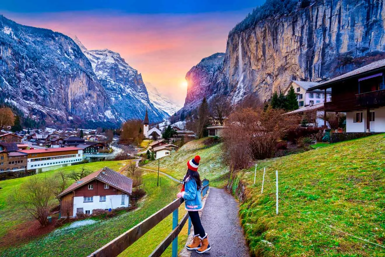 Person standing on a path overlooking a village nestled in a valley with mountains, snow-capped peaks, and a vivid sunset sky.