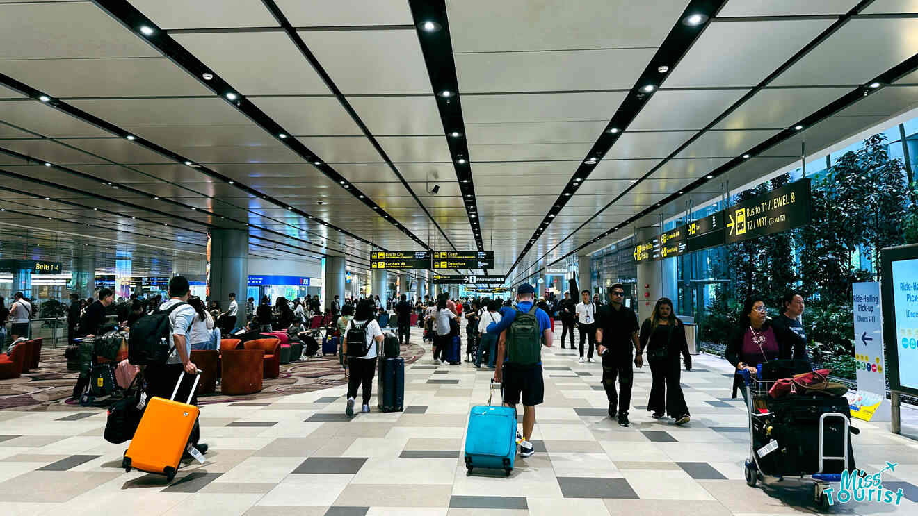 A busy airport terminal with travelers pulling luggage. Overhead signs direct to gates and amenities. People are gathered and walking around under the brightly lit ceiling.