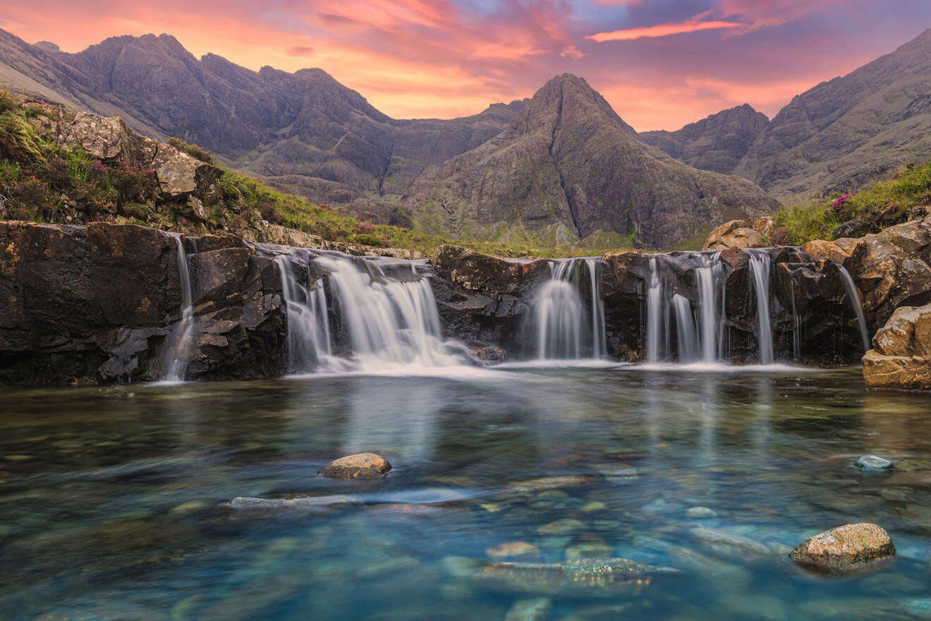 A clear mountain stream cascades over rocks under a vibrant sunset sky, with jagged peaks in the background.