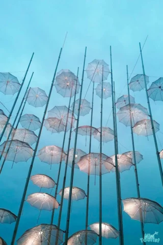 A group of transparent umbrellas are suspended on poles against a cloudy blue sky, some illuminated with small lights.