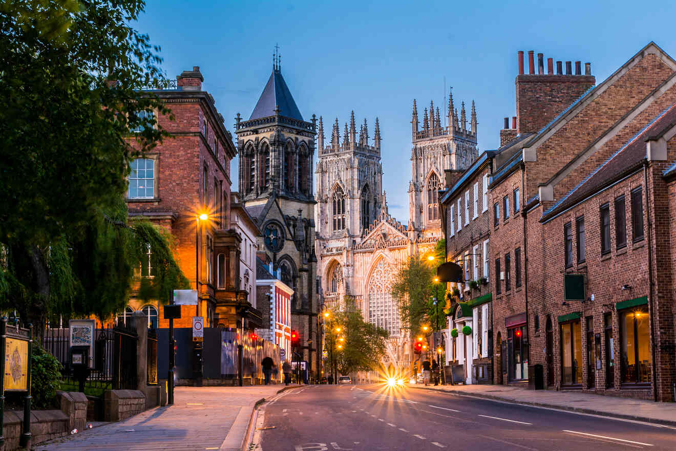 Street view of York, England at dusk, with York Minster cathedral illuminated in the background and old brick buildings lining the quiet road.