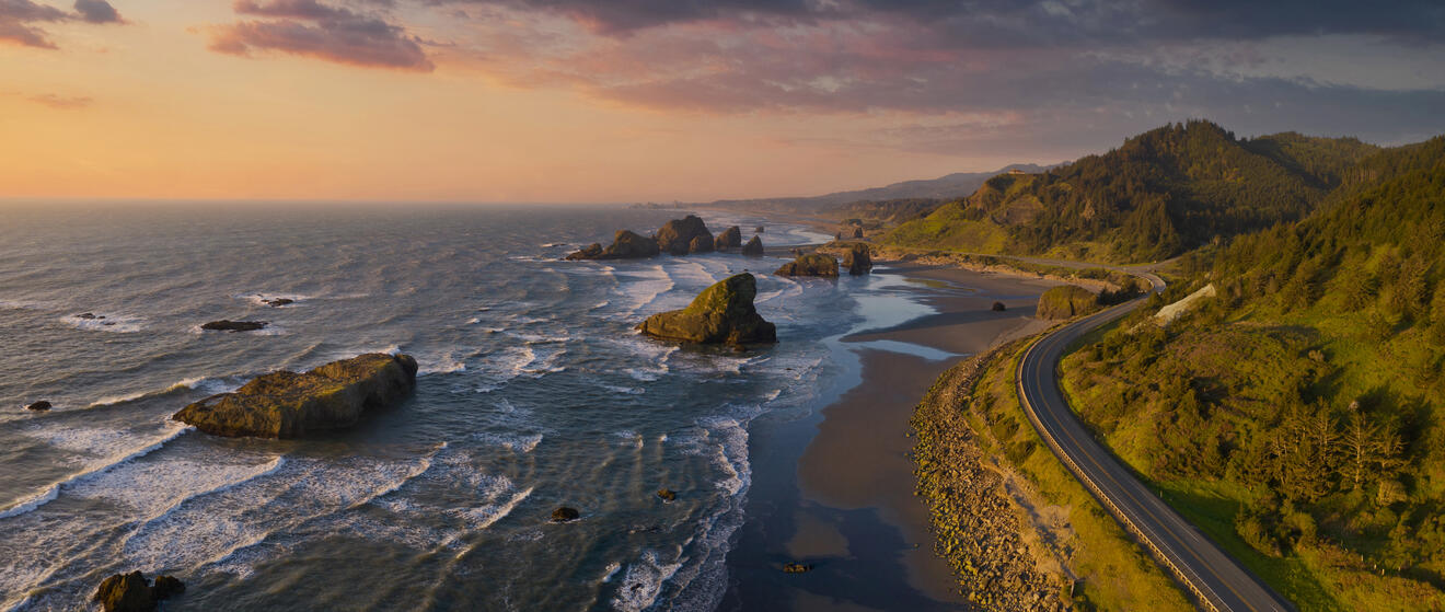 A coastal landscape featuring rugged cliffs, waves crashing against rock formations, and a winding road parallel to the shoreline during sunset.